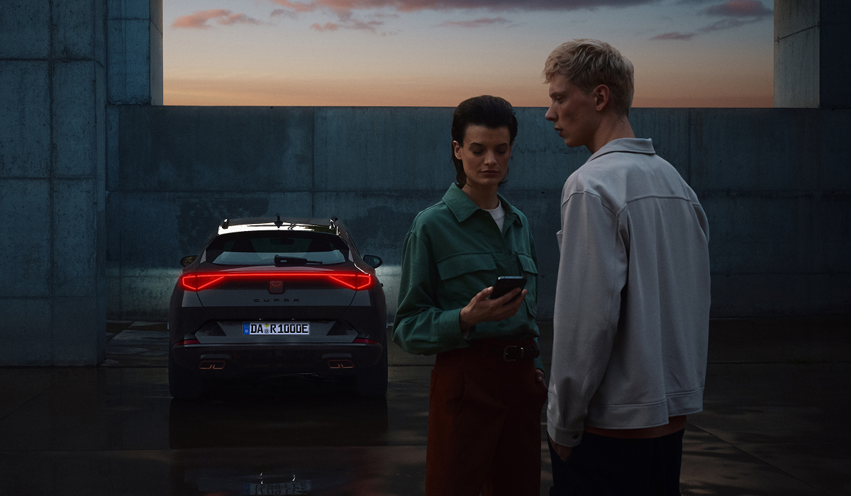 Zwei Personen neben einem CUPRA Formentor in Heckansicht bei Abendstimmung mit leuchtendem Rücklicht eine Person hält ein Smartphone